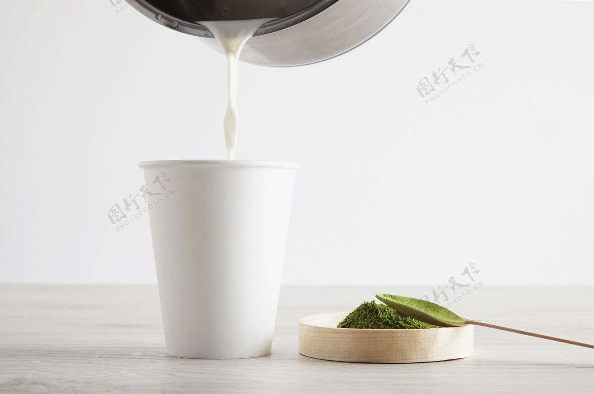 桌子侧视图：木制桌上的白色纸杯和优质有机日本抹茶 为现代拿铁咖啡的制作做好准备演示第三步将一点热牛奶倒进杯子里木材饮料抹茶