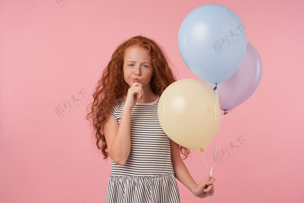 雀斑迷人的红发卷发长发女孩穿着休闲服站在粉色背景下 若有所思地看着镜头 举起手托着下巴 手里拿着气球有色折叠孩子