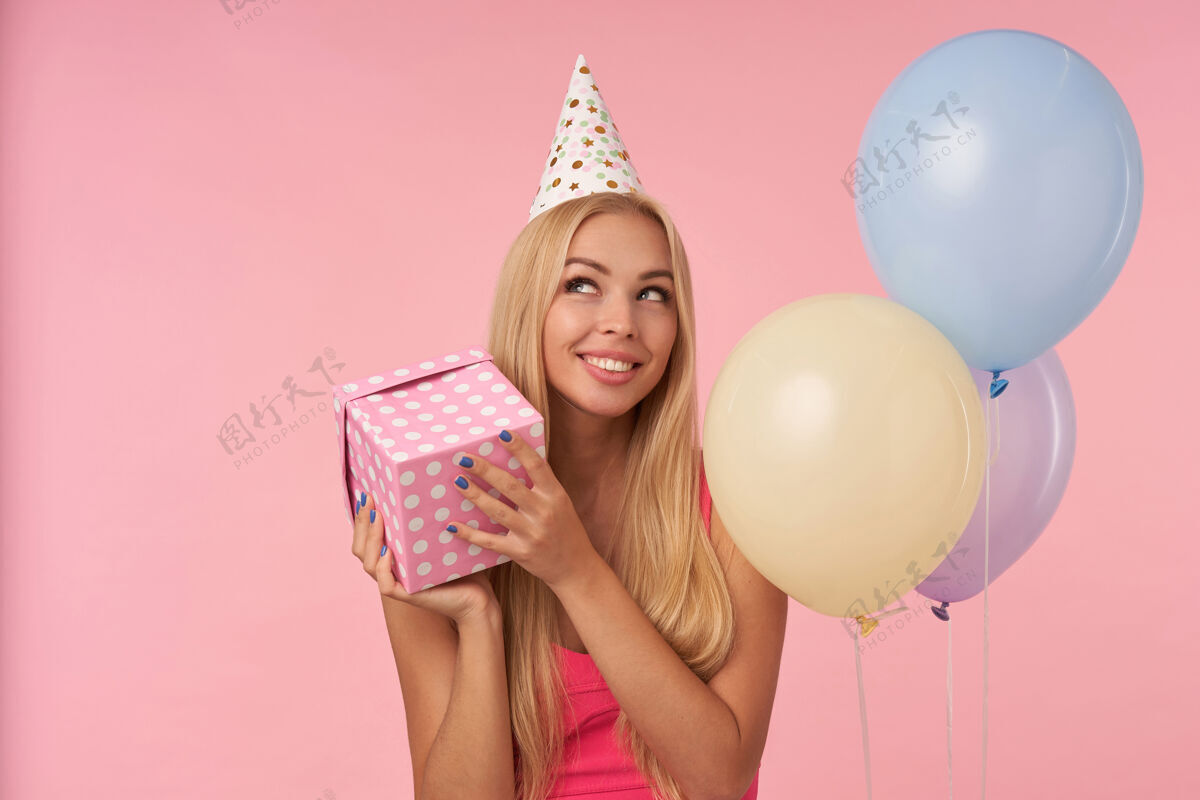 情感身着粉色上衣 头戴生日帽的快乐金发女郎的肖像 在五彩气球中摆姿势 开心地看着一边 手里拿着礼物盒 站在粉色背景上微笑肖像欢快