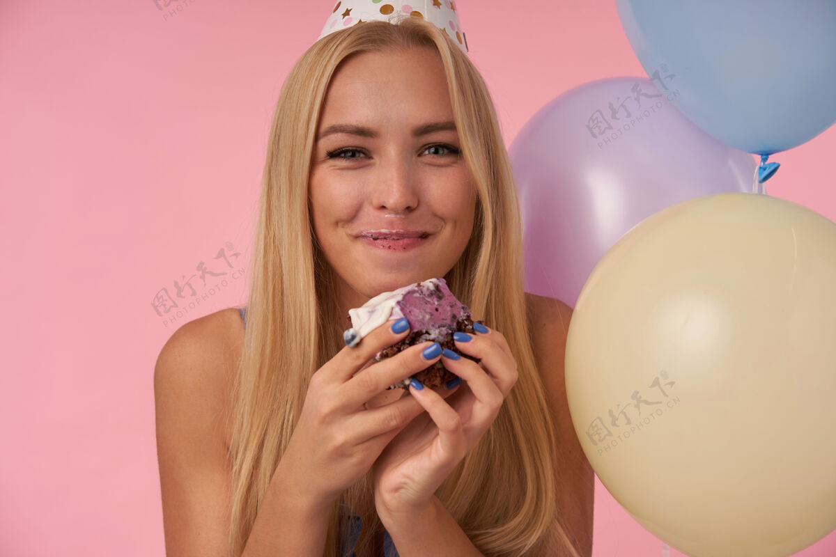摆姿势快乐的年轻漂亮的金发女郎在五颜六色的气球里摆着姿势 嘴里塞满了节日蛋糕 开心地看着镜头 笑容满面 在粉色的背景下展现出她愉悦的情感享受女性年轻