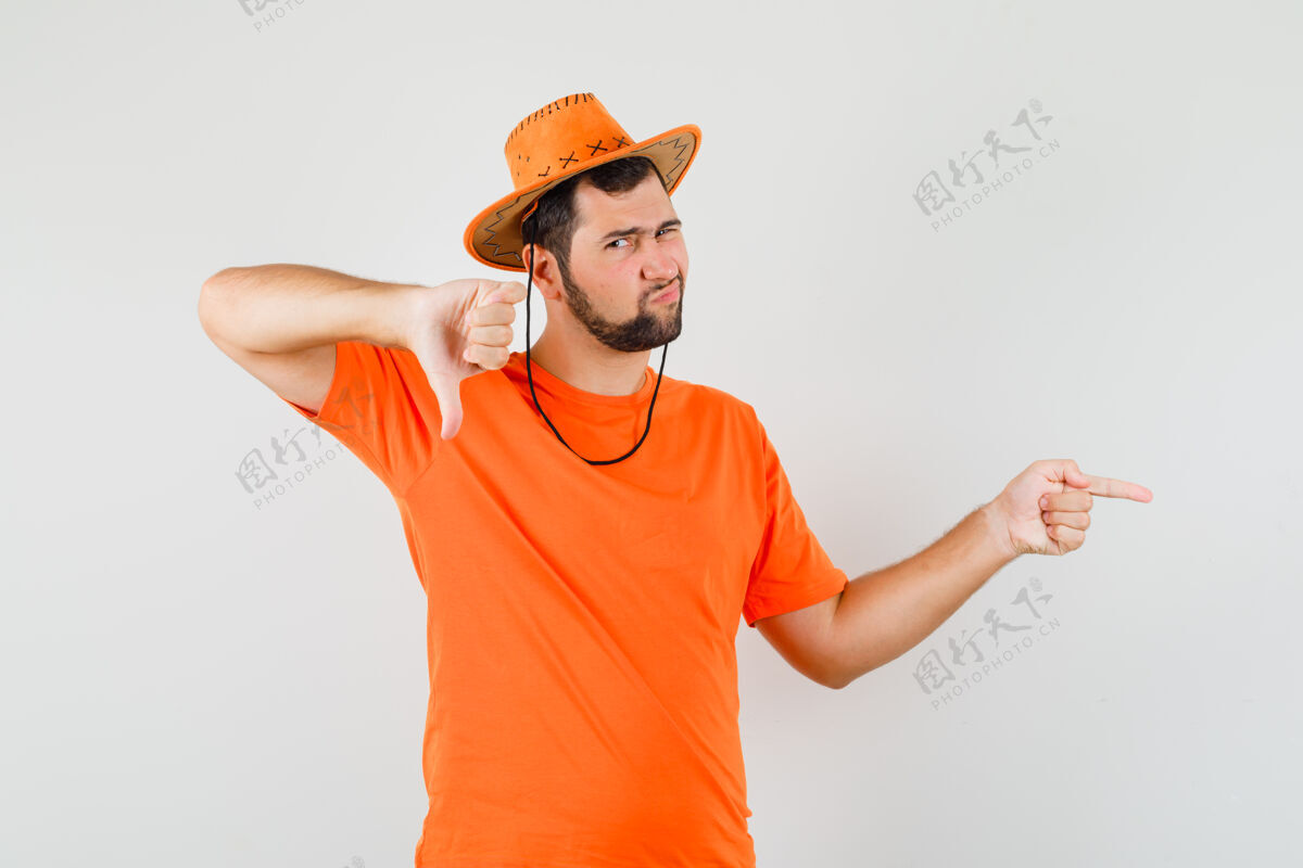 年轻年轻人指着旁边 大拇指朝下 穿着橙色t恤 戴着帽子 看上去很不满意正视图时尚休闲手势