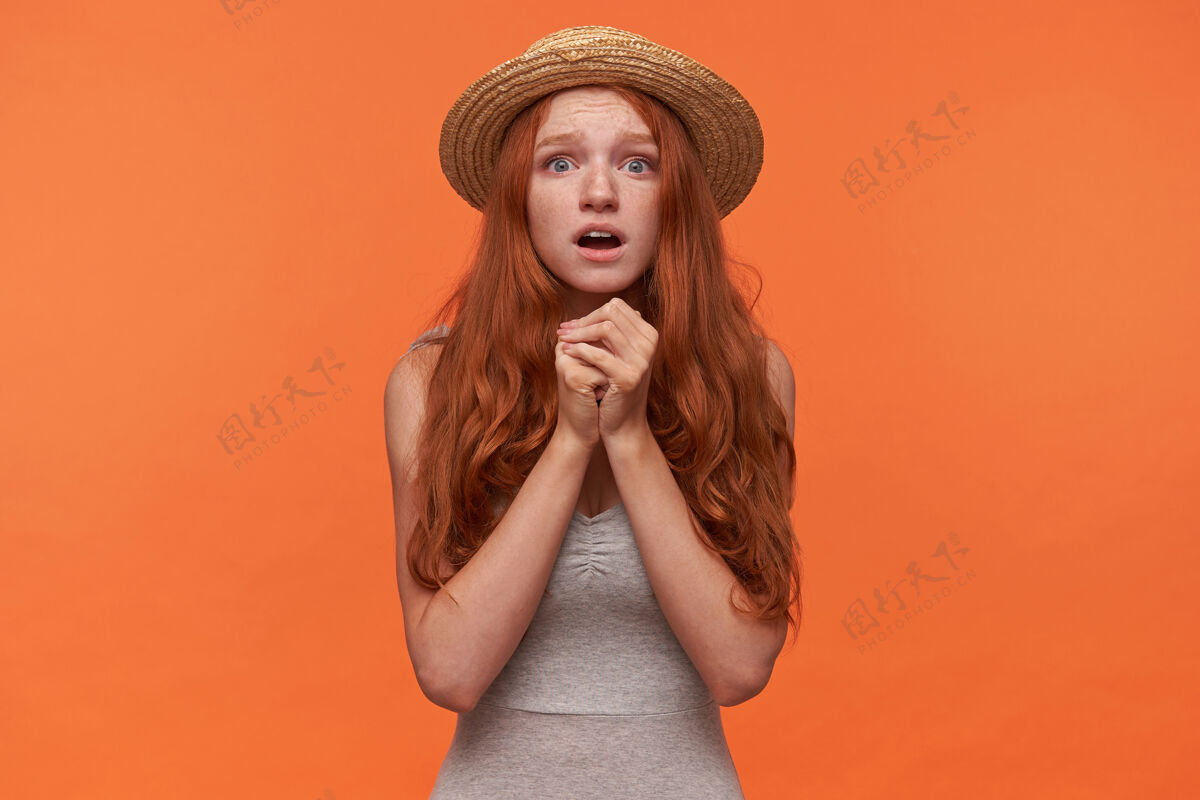 波浪一幅绝望的年轻红发孤独女性的肖像 身着灰色衬衫 头戴水手帽 双手举在一起看着相机 希望能解决她的问题 孤立在橙色背景下20多岁头发衬衫