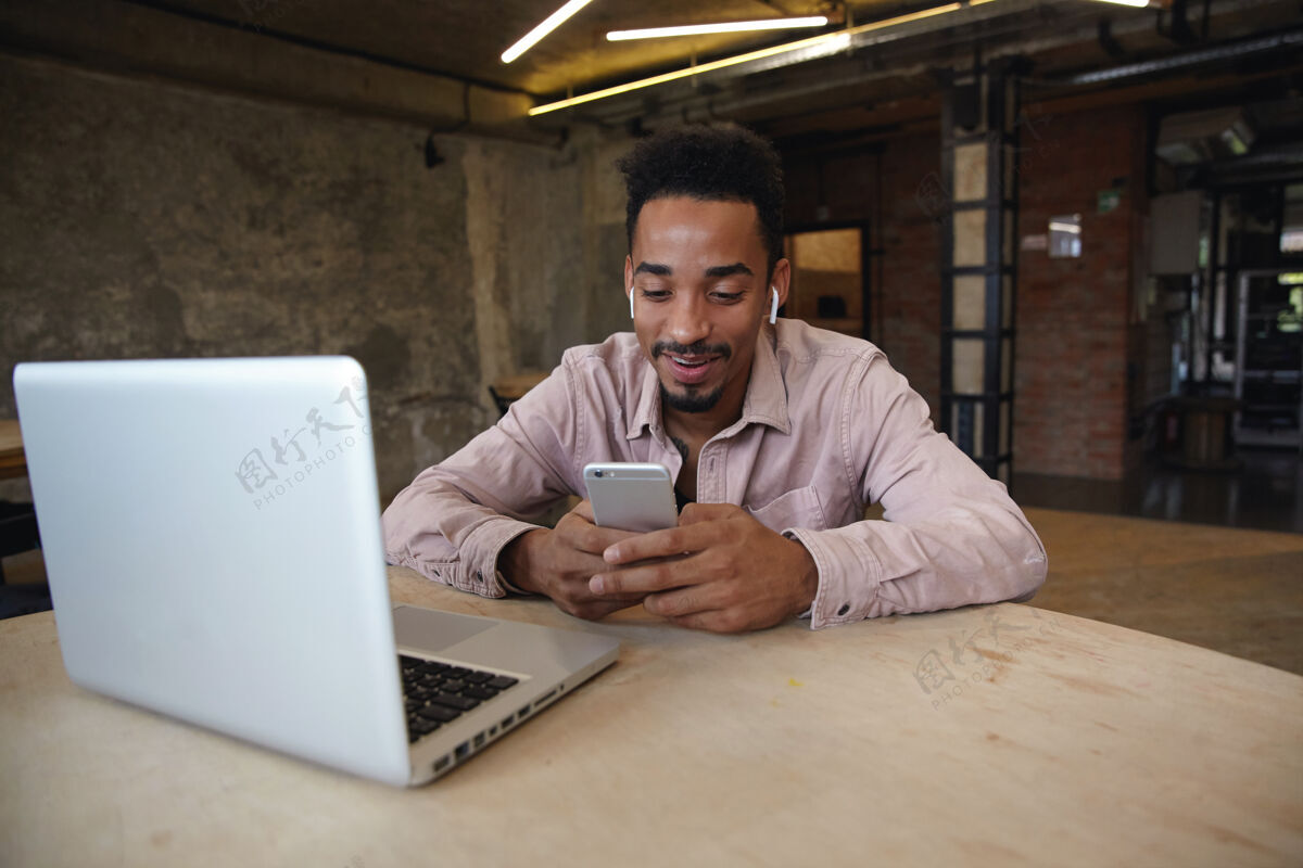 远程正面帅气的留着黑胡子的男人 穿着米色衬衫 在同事的空间里远程工作 手里拿着手机 查看社交网络开朗室内商人