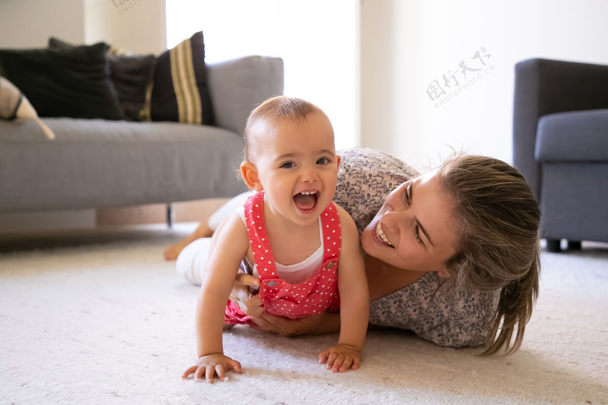 婴儿快乐的妈妈和小女孩躺在客厅的地毯上快乐的金发妈妈和快乐的女儿在家里玩耍 微笑着可爱的宝宝张着嘴笑着母性和周末的概念玩地毯躺着