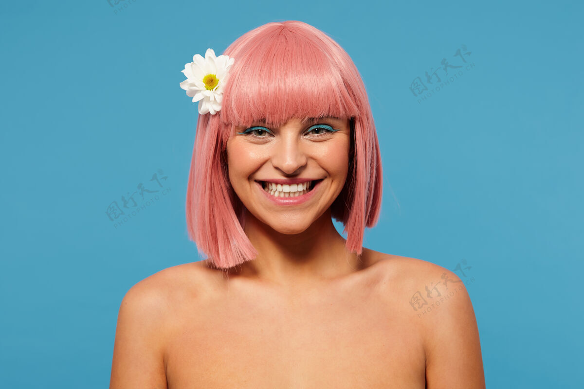湛蓝快乐的年轻漂亮的粉色头发的女人 留着鲍勃式的发型 愉快地看着镜头 带着迷人的笑容 站在蓝色背景下 头发上插着白花优雅开朗肩膀
