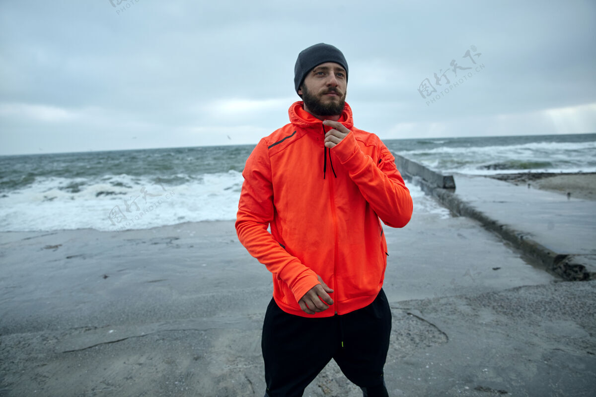 户外在寒冷的清晨 年轻的运动型深色头发 蓄着浓密胡须的男子在海边摆姿势 准备每天的锻炼 面容平静地向前看健康暴风雨运动员