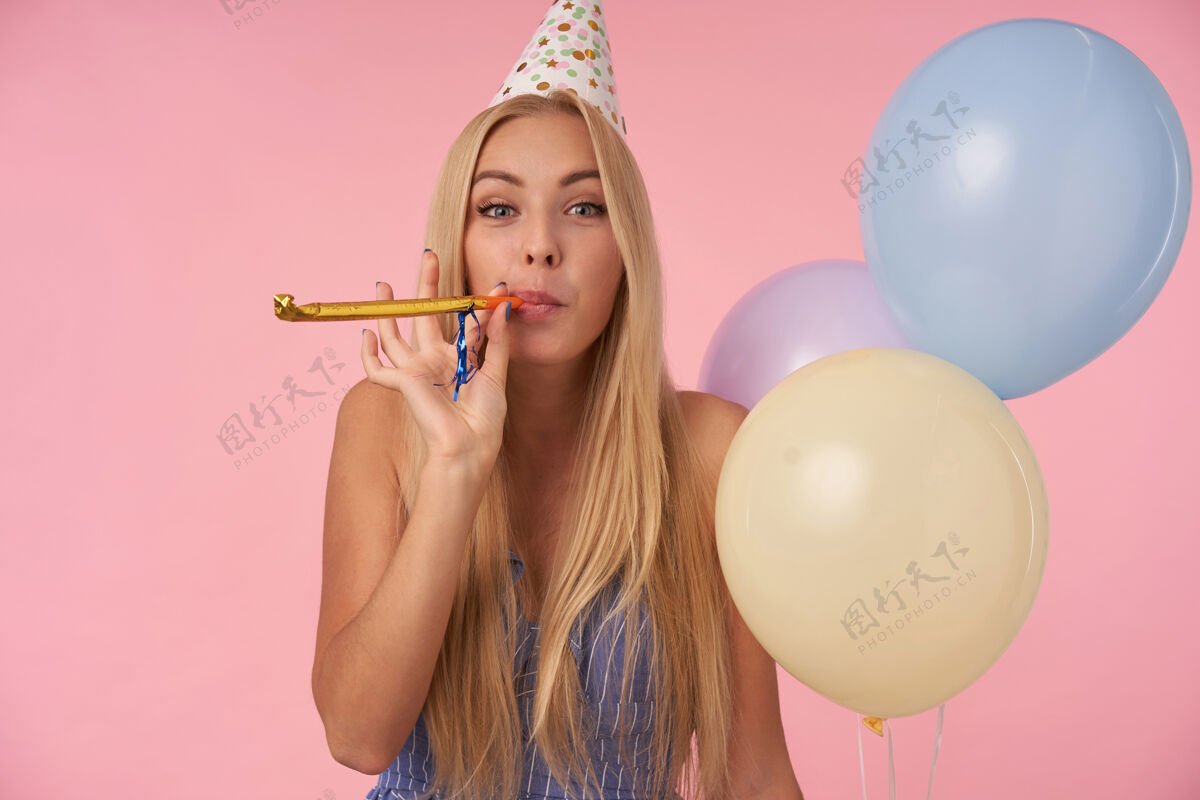 金发快乐的长发金发女人 穿着蓝色夏装 戴着生日帽 手里拿着一束氦气球 兴高采烈地看着相机 吹着派对号角 孤立在粉色背景下正面长发迷人