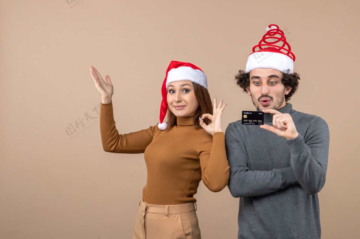 红色圣诞节心情激动满意酷情侣戴着红色圣诞老人帽展示银行卡 女人做ok手势穿着心情快乐