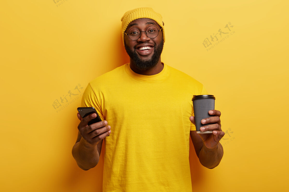 无线穿着黄色衣服的快乐黑人的孤立镜头 输入信息 在智能手机上下载新应用程序 从一次性杯子里喝咖啡 有着牙齿般的微笑 洁白的牙齿 浓密的鬃毛设备牙齿杯子