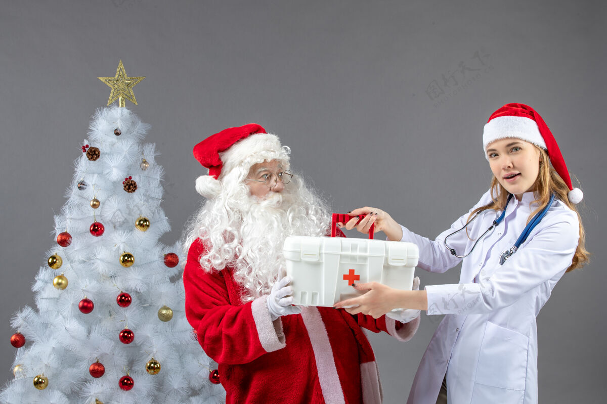 圣诞老人圣诞老人和女医生的前视图 女医生在灰色的墙上拿着急救箱庆祝第一快乐