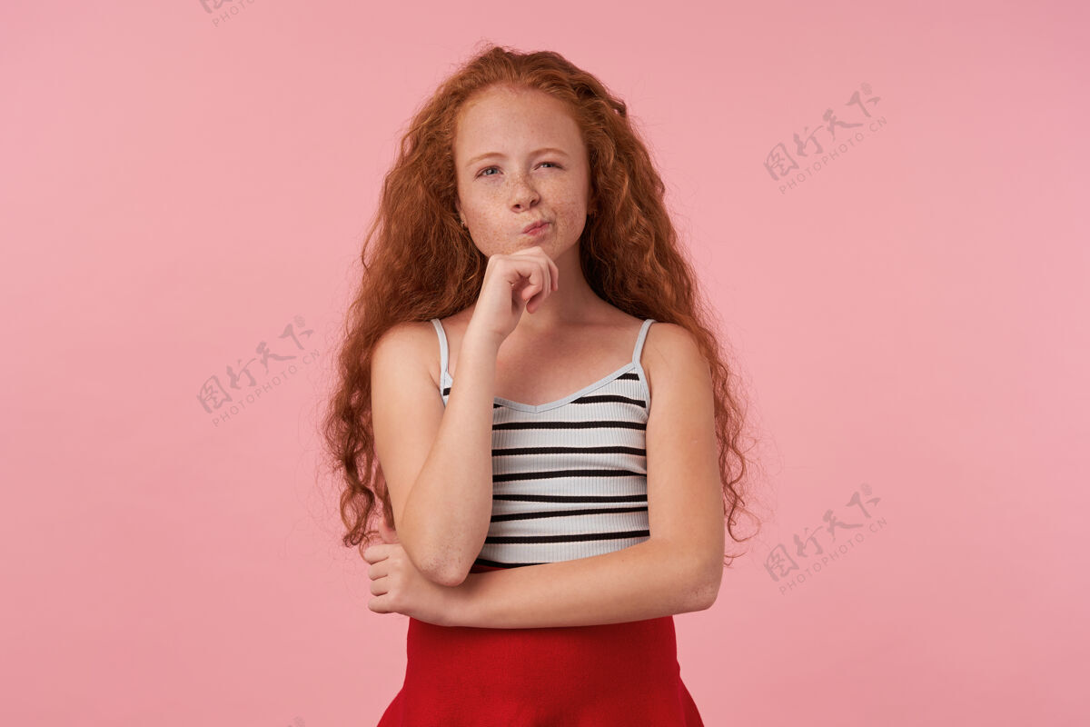 孩子室内拍摄的红发卷发女童子 休闲发型 穿着红色裙子和条纹上衣 抬起手托着下巴 若有所思地看着相机 隔离在粉色背景下孩子体贴折叠