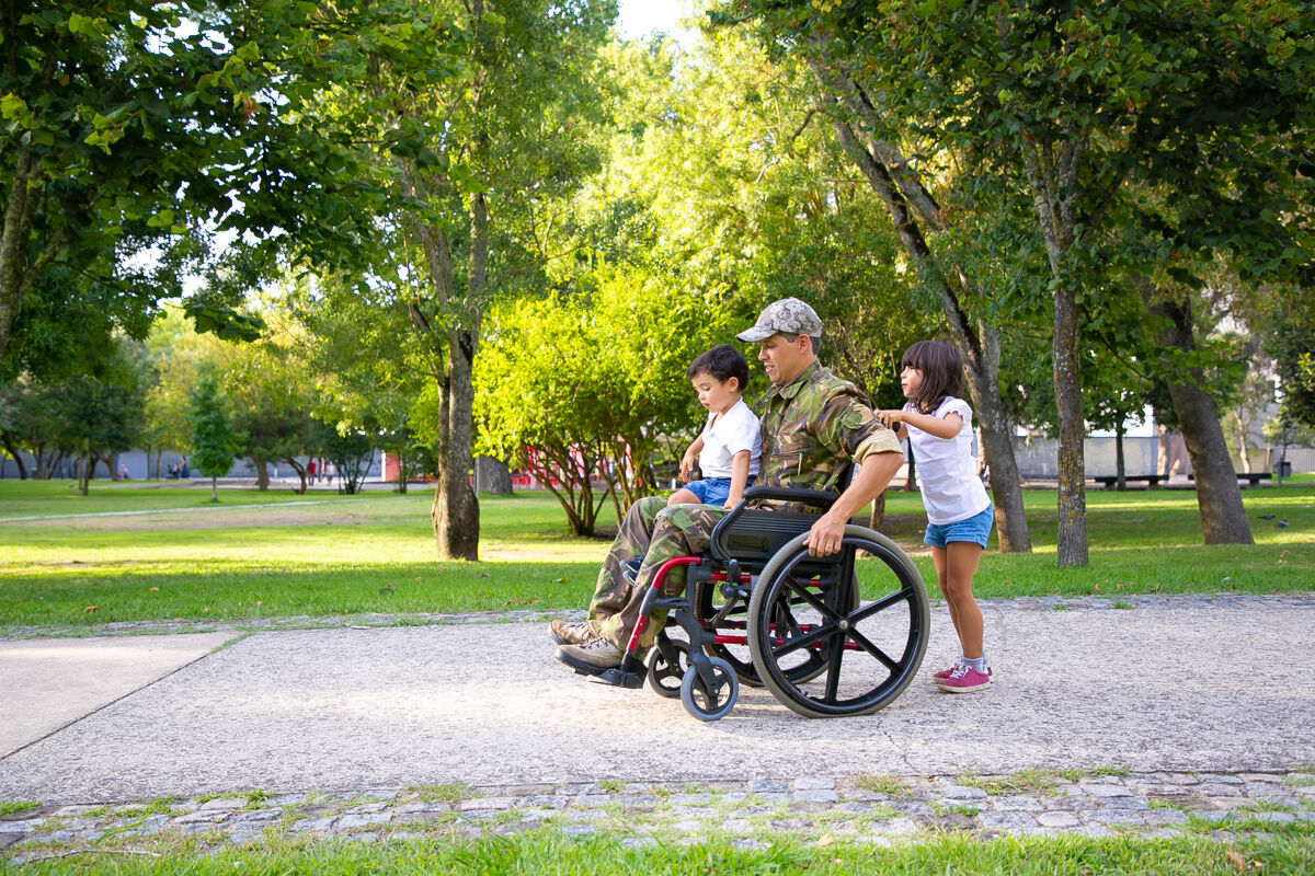 肖像残疾退伍军人和两个孩子在公园散步男孩坐在爸爸的腿上 女孩推着轮椅退伍军人的战争或残疾的概念军队孩子残疾人