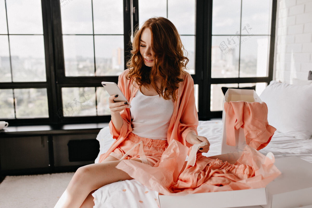 睡衣可爱的女孩在卧室里读着电话留言快乐的微笑的女人拿着电话坐在床上人快乐休闲