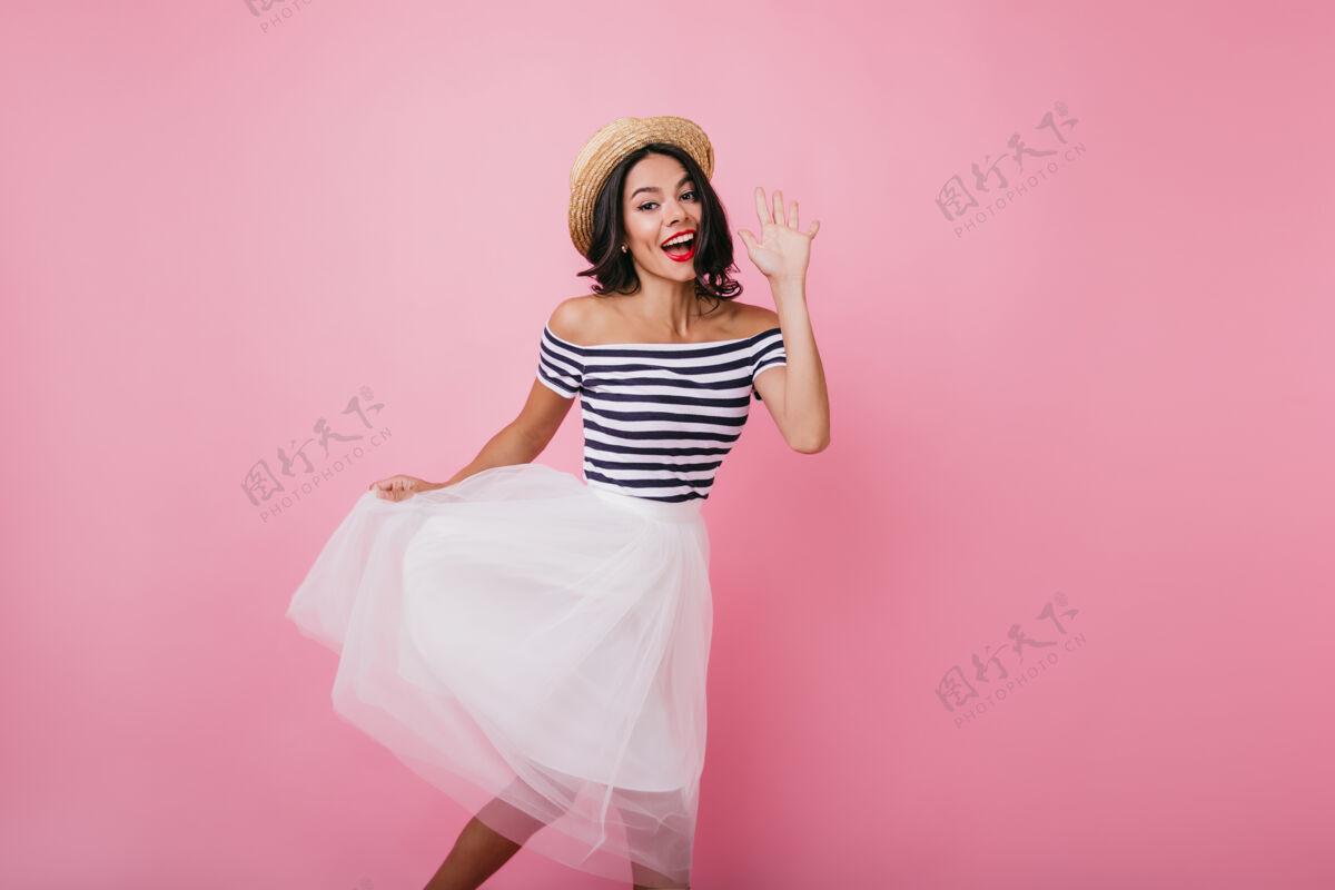舞者幸福的棕褐色女人一边跳舞一边挥舞着手室内画像中 喜气洋洋的黑发女孩穿着优雅的郁郁葱葱的裙子裙子帽子积极