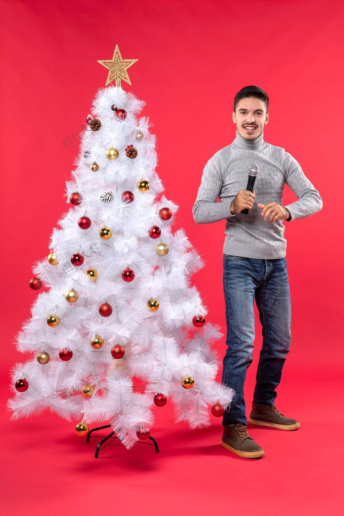 自信圣诞节的心情与自信的家伙穿着牛仔裤站在装饰圣诞树附近手持麦克风 愉快地说人男人装饰