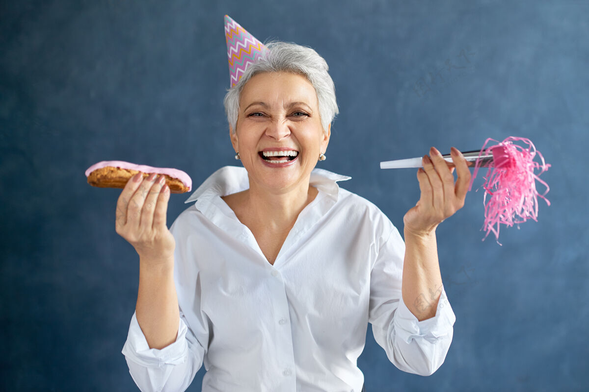 可爱一头白发的成熟女性的肖像 戴着粉红色的派对帽 手持哨子和喇叭吹退休女士