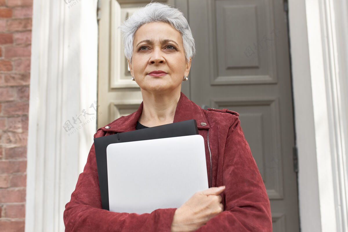 成人时尚的50岁白人妇女手持便携式电脑在屋外摆姿势的画像退休行政人员电脑