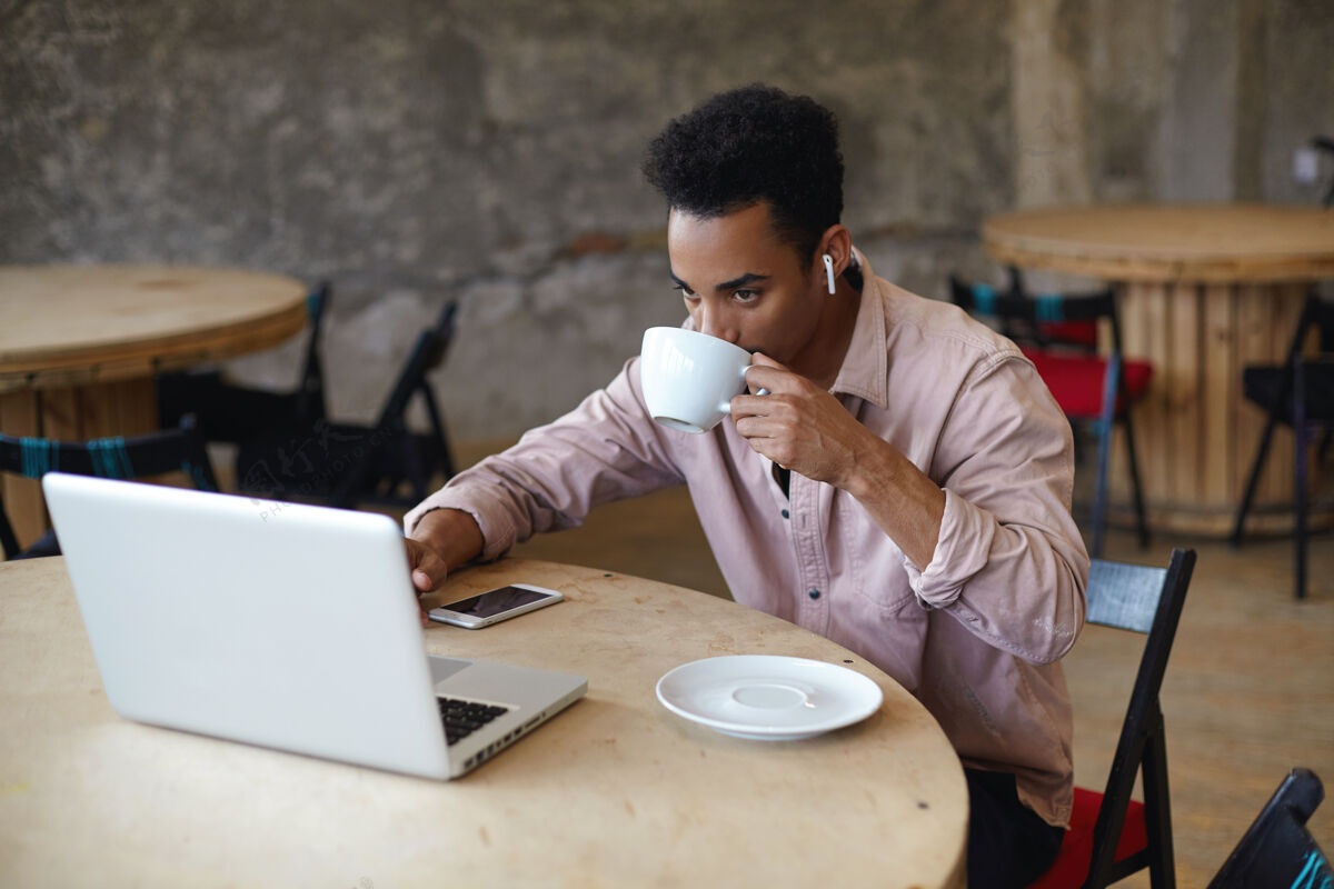 肖像年轻的留着胡须 皮肤黝黑 留着短发的商人坐在米色衬衫的木制圆桌旁 一边在城市咖啡馆里喝咖啡 一边用笔记本电脑准备与客户见面的材料不留胡子耳机摆姿势