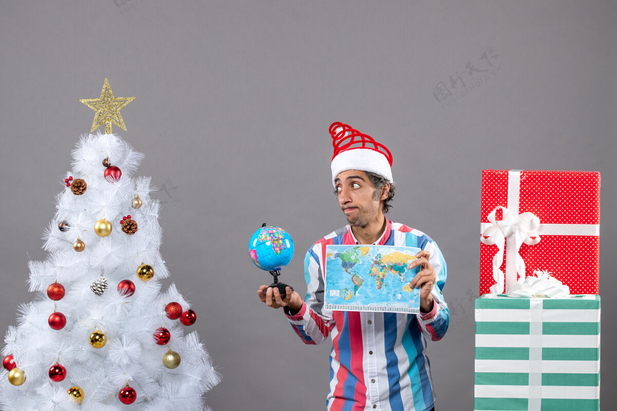 家伙前视图沮丧的男子与螺旋弹簧圣诞帽举行世界地图和地球仪地图灰色圣诞树