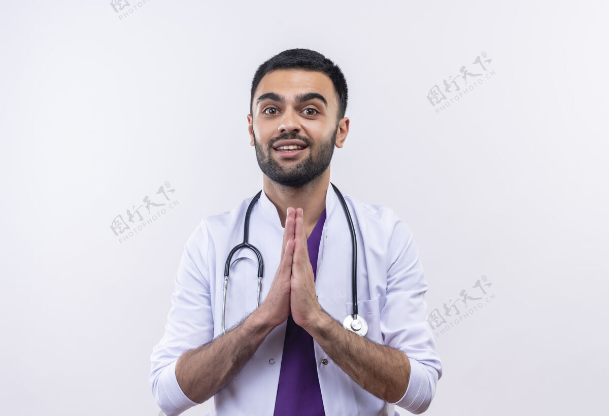 展示微笑着的年轻男医生穿着听诊器医用长袍 在孤立的白色背景上展示祈祷的姿势年轻医疗白色
