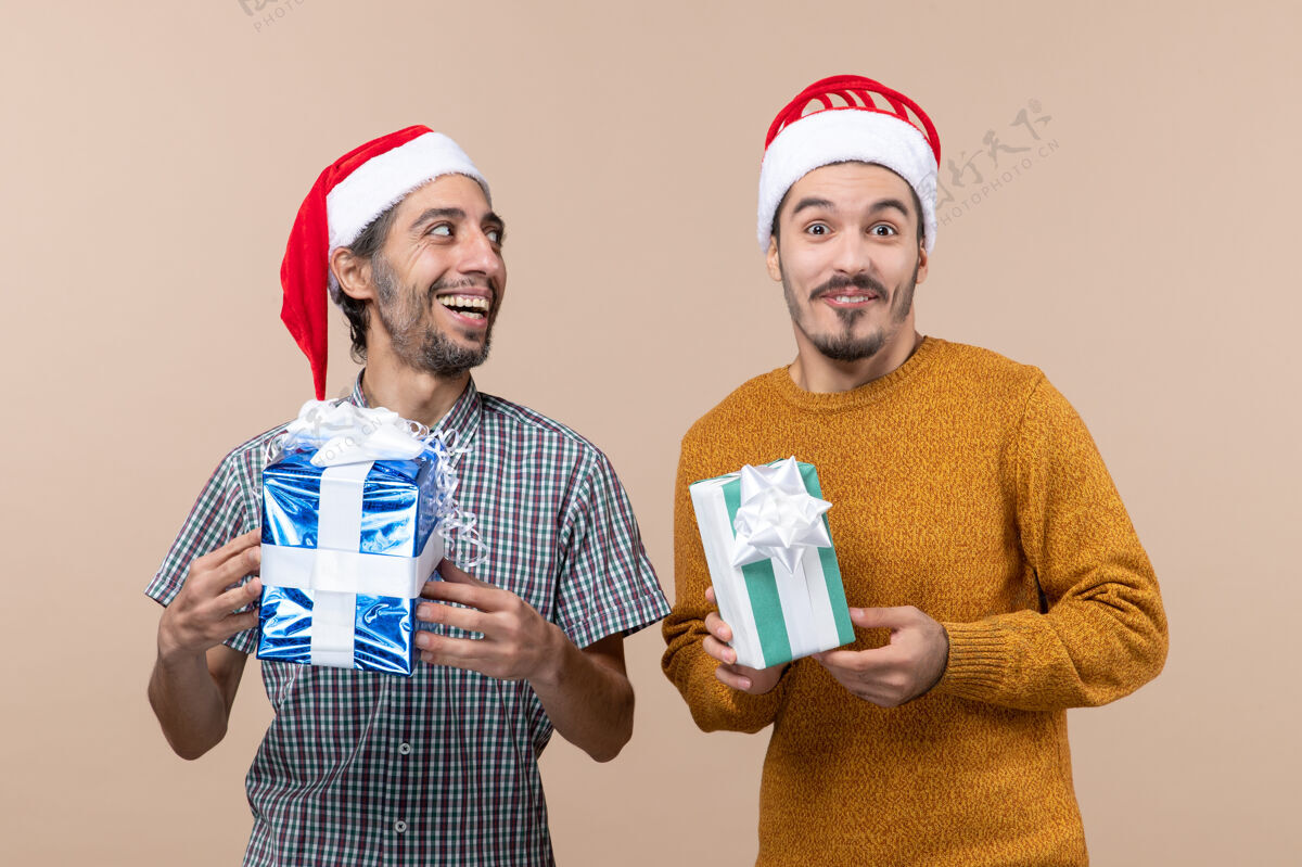 快乐正面图：两个戴着圣诞帽 拿着圣诞礼物的快乐男人 背景是米色的男人两个两个快乐的男人