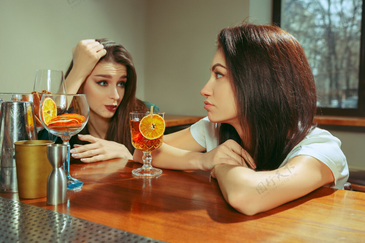 朋友女朋友在酒吧喝酒她们坐在一张摆着鸡尾酒的木桌旁她们穿着休闲服白天友谊交流