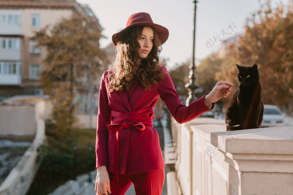阳光美丽时尚的女人穿着紫色西装在城市街头 春夏秋冬时节时尚潮流戴着帽子 看着一只猫秋天优雅漂亮