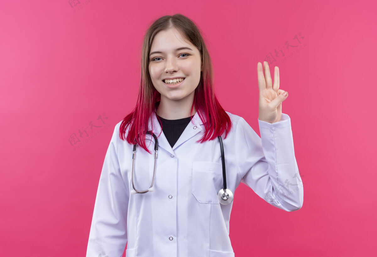 微笑带着微笑的年轻医生女孩穿着听诊器医用长袍 在粉色孤立的背景上显示三个展示穿着长袍
