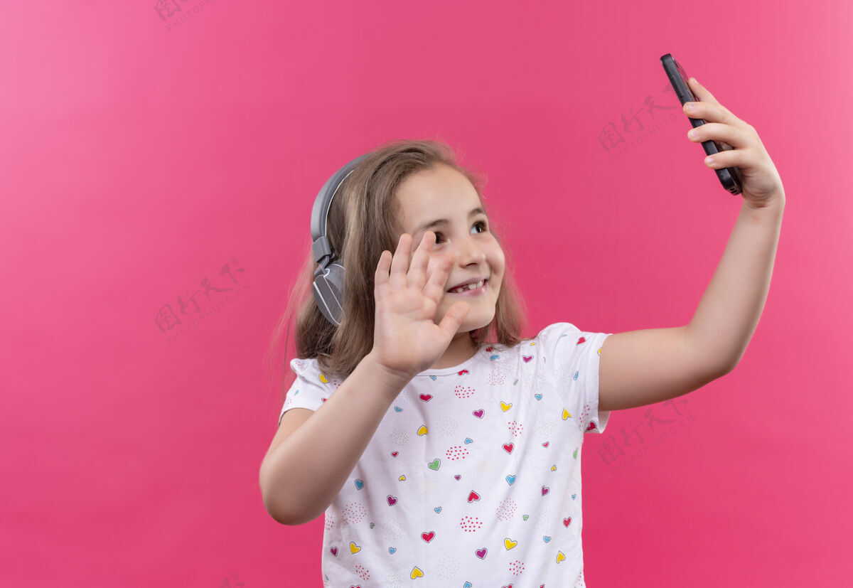 学校微笑的小女孩穿着白色t恤 戴着耳机 在孤立的粉色背景下讲电话女孩电话小