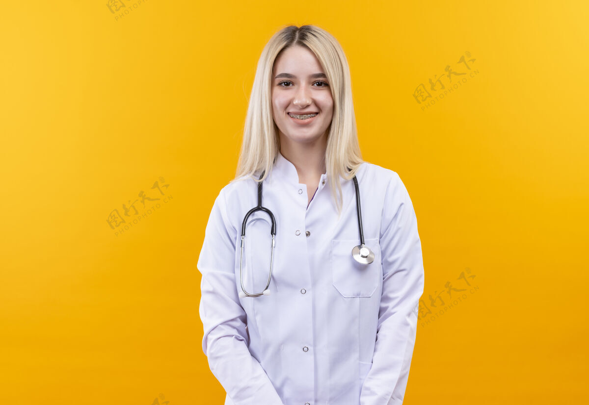 微笑微笑的医生年轻女孩穿着医用长袍戴着听诊器和牙套在孤立的黄色背景上黄色背带女孩
