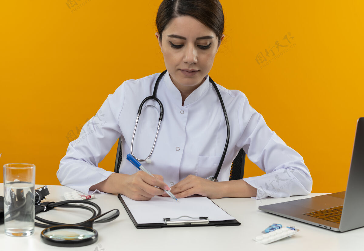 工作年轻的女医生穿着医用长袍带着听诊器坐在办公桌上用电脑和医疗工具在剪贴板上写隔离黄色背景穿着电脑办公桌