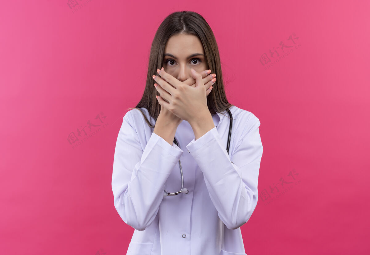 女孩被吓坏了的年轻女医生穿着听诊器医用长袍捂住嘴 背景是孤立的粉红色罩子医生粉红