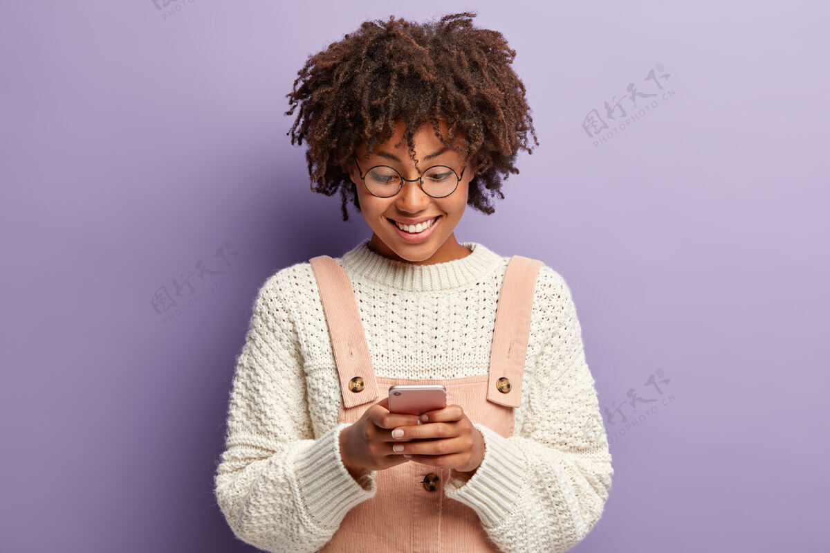小玩意快乐卷发女人的室内镜头 脸上洋溢着喜悦的表情 读着男友的美好留言 专注于智能手机互联网移动工作服