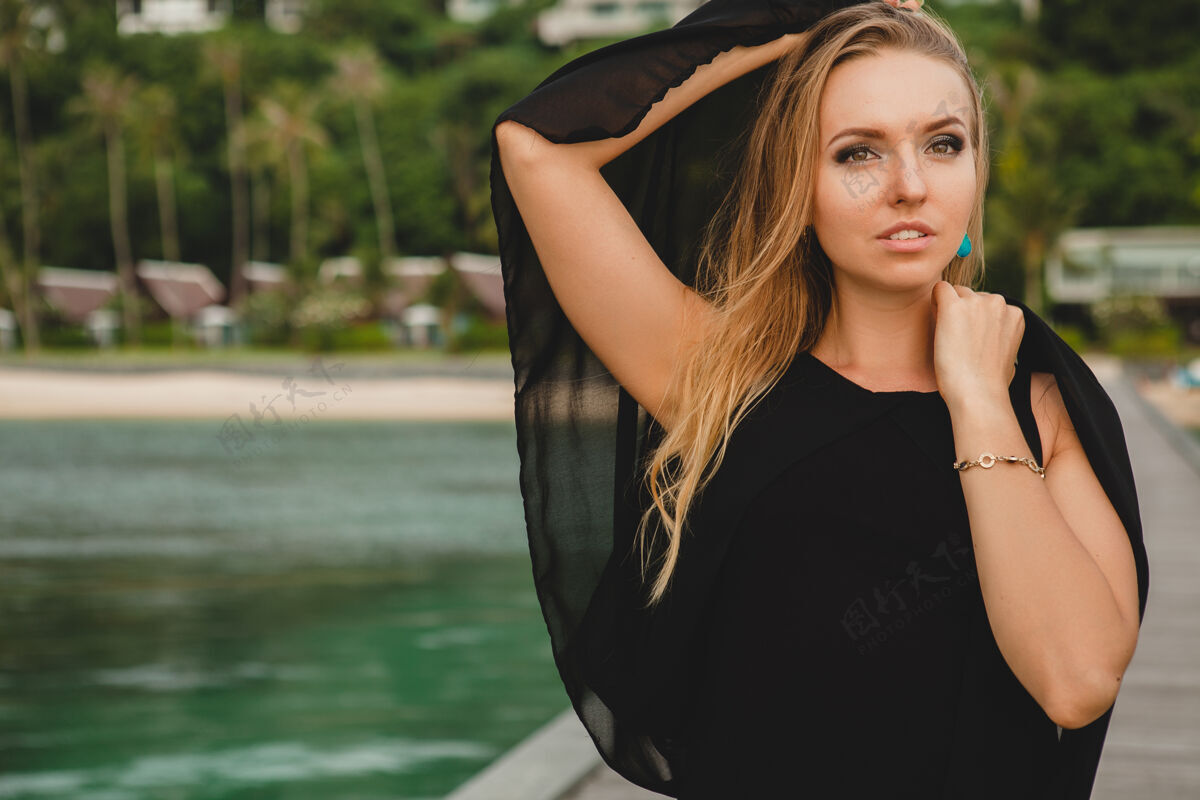 摆姿势穿着黑色连衣裙的美丽迷人的女人在豪华度假酒店的码头上摆姿势 暑假 热带海滩热带模特头发