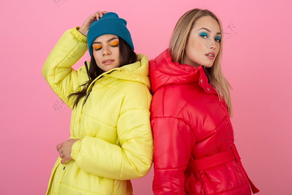 女孩两个迷人的女孩在粉红色的背景下 穿着鲜艳的红色和黄色的彩色冬季羽绒服摆姿势外套蓝色时尚