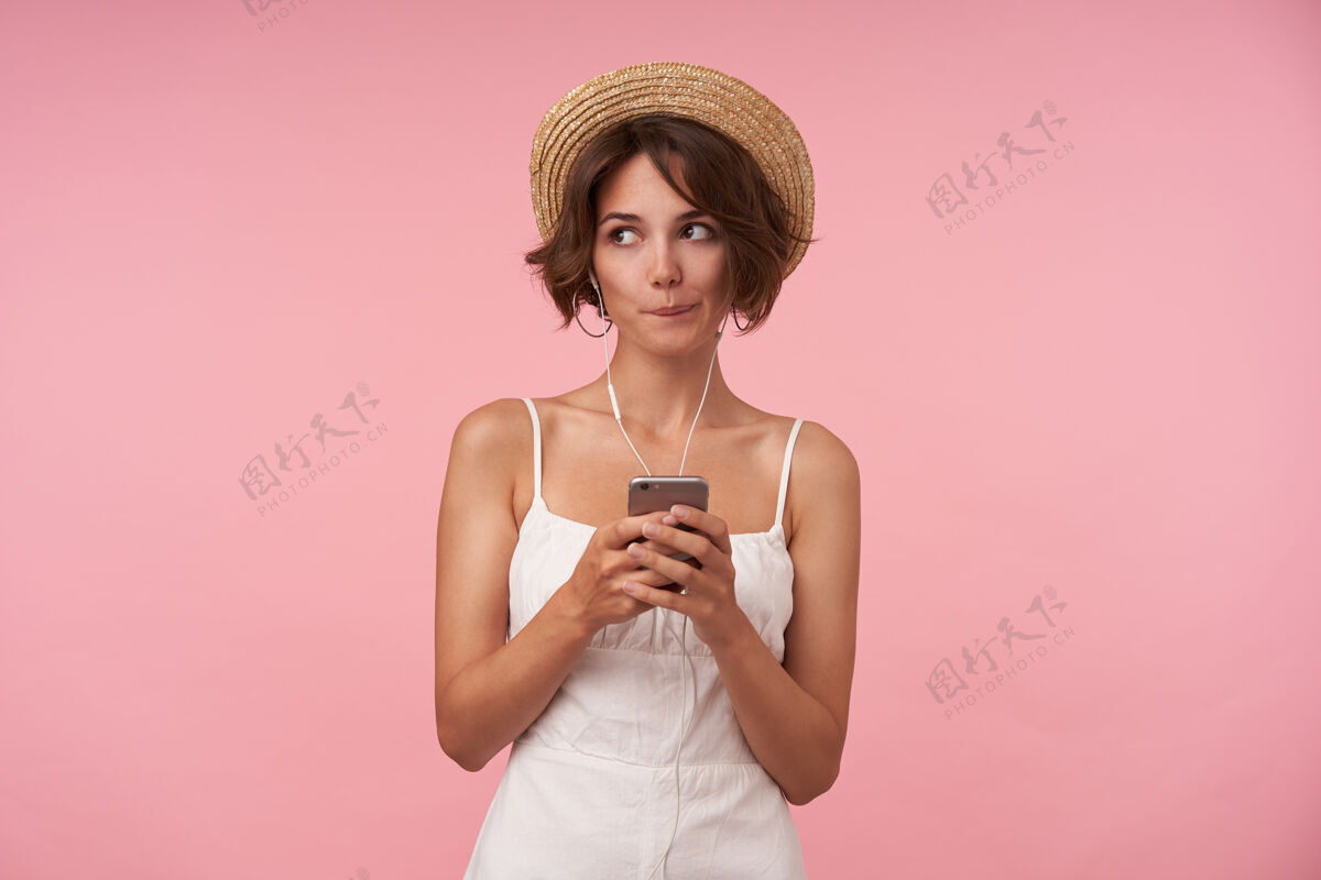 理发困惑的年轻黑发女子一边看着一边 一边咬着内衣 一边举起手拿着手机 身穿白色夏装 头戴草帽美丽沉思女士