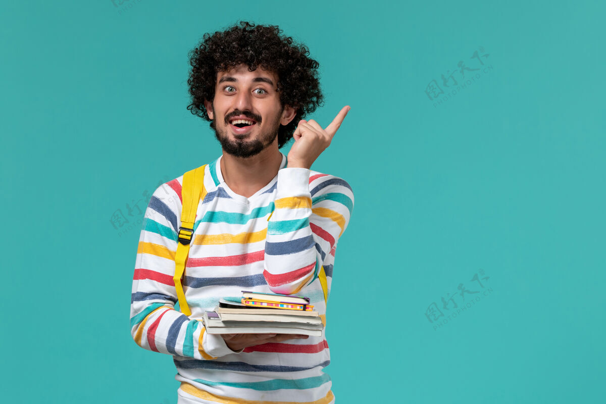 非洲身穿条纹衬衫 背着黄色背包 在蓝色墙上拿着书的男学生的正视图大学正面学生