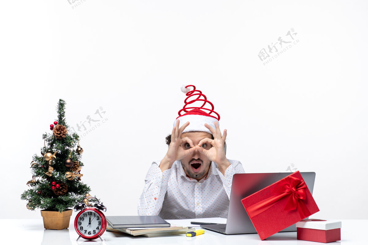 圣诞老人在白色背景的办公室里 年轻的商人戴着滑稽的圣诞老人帽子 在圣诞树上做着戴眼镜的手势 这让人大吃一惊圣诞节圣诞老人商人