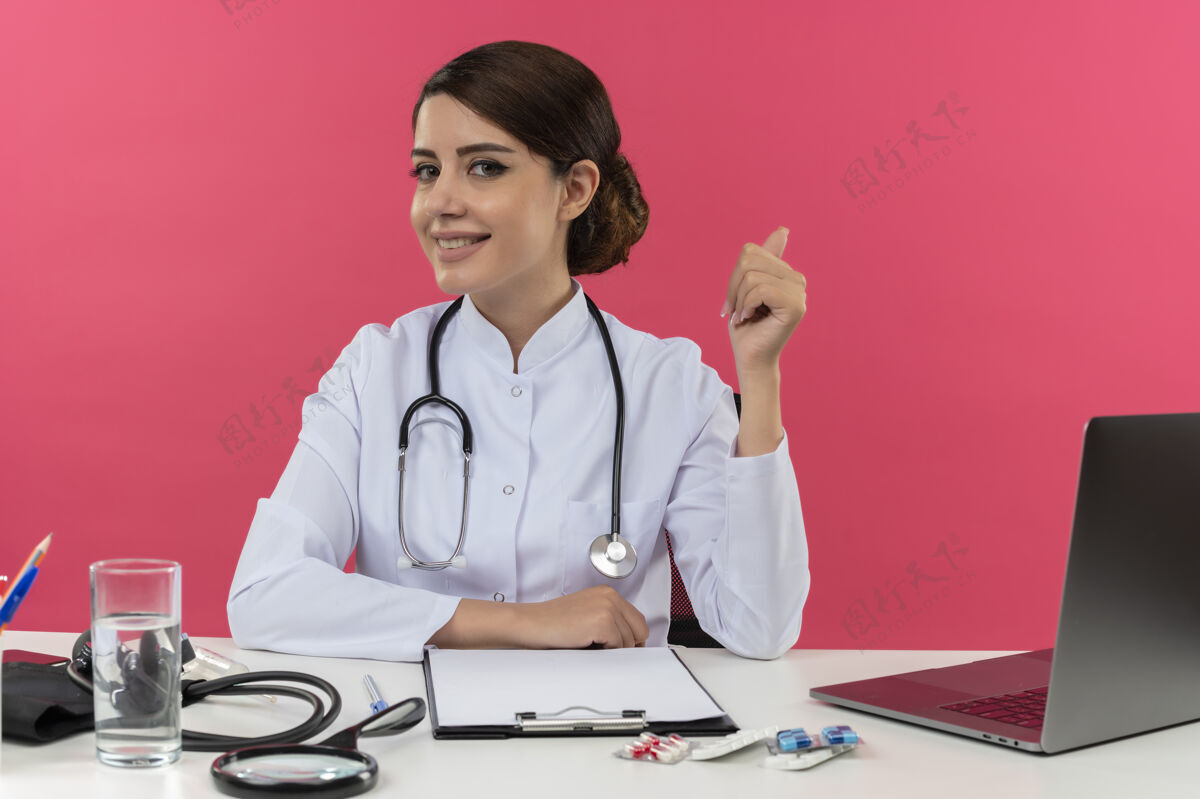 医生面带微笑的年轻女医生穿着医用长袍 手持听诊器坐在办公桌旁 拿着医疗工具在电脑上工作 她竖起大拇指放着复印空间工作坐着工具
