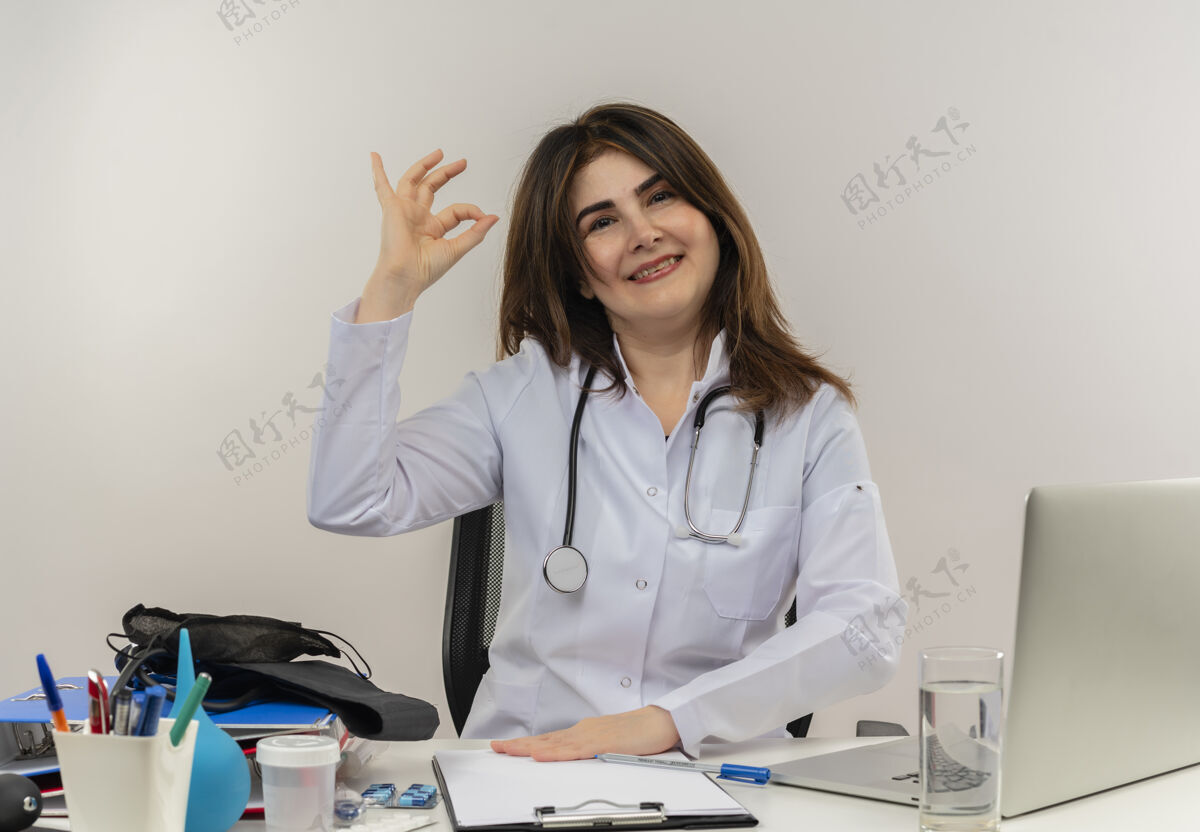 好的微笑的中年女医生 穿着医用长袍 带听诊器 坐在办公桌旁 用笔记本电脑和医疗工具 在白色背景上显示出良好的姿势 并留有复印空间展示笔记本电脑中年