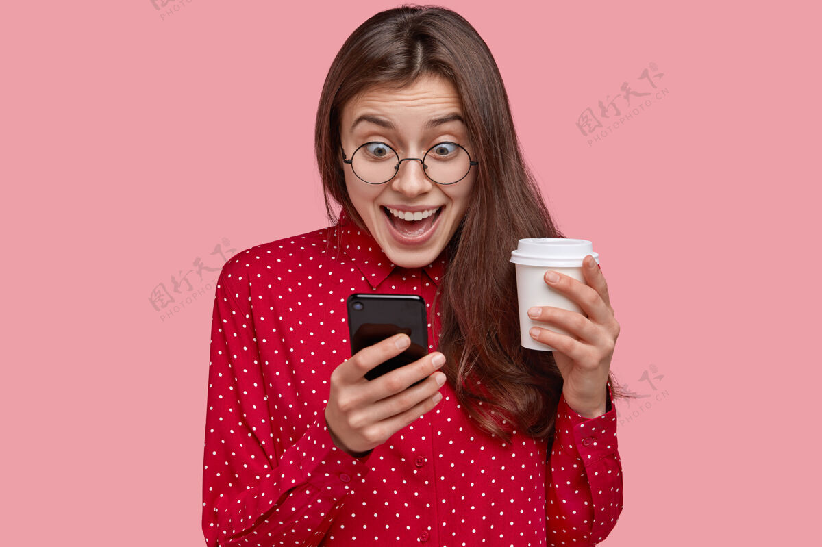 设备照片中的年轻女子兴高采烈地查看社交网络 玩手机游戏 浏览网络应用程序 喝外卖咖啡 穿着红色衣服惊奇眼镜眼镜