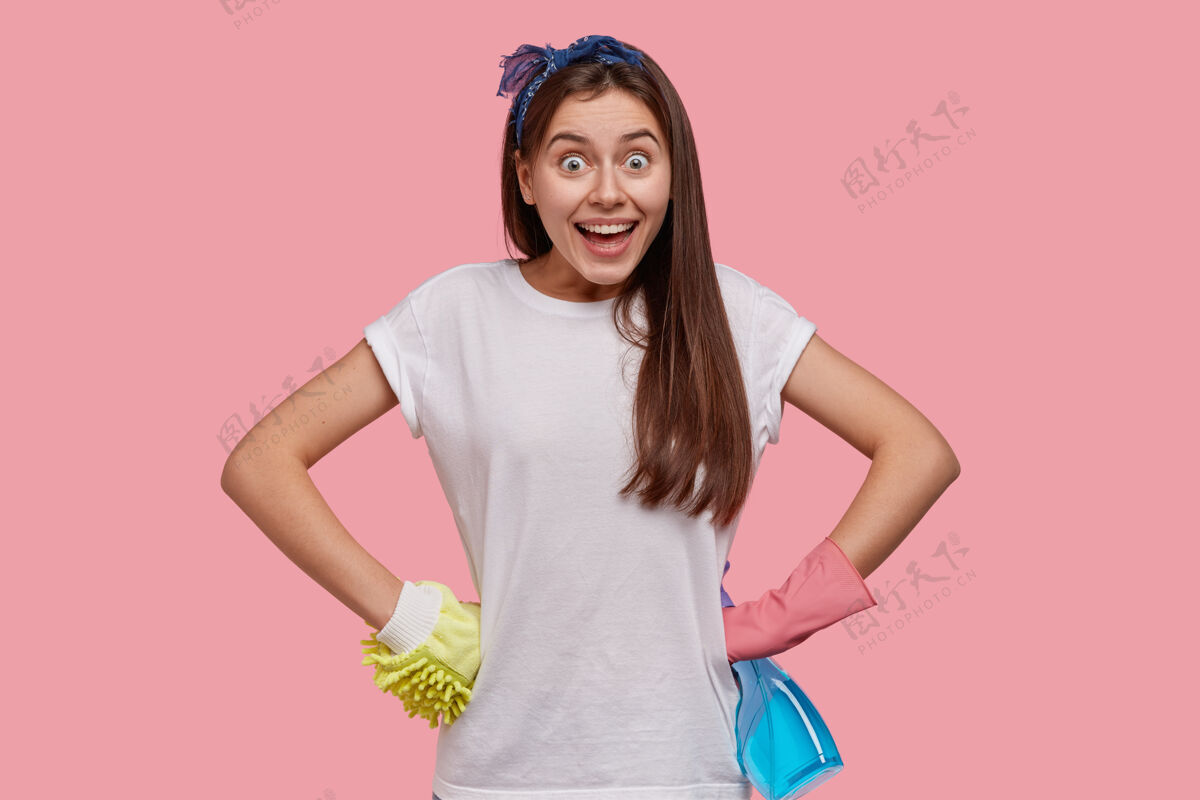 摆姿势照片中的快乐年轻女佣手放在腰上 穿着休闲的白色t恤 头带 防护手套满意清洁清洁