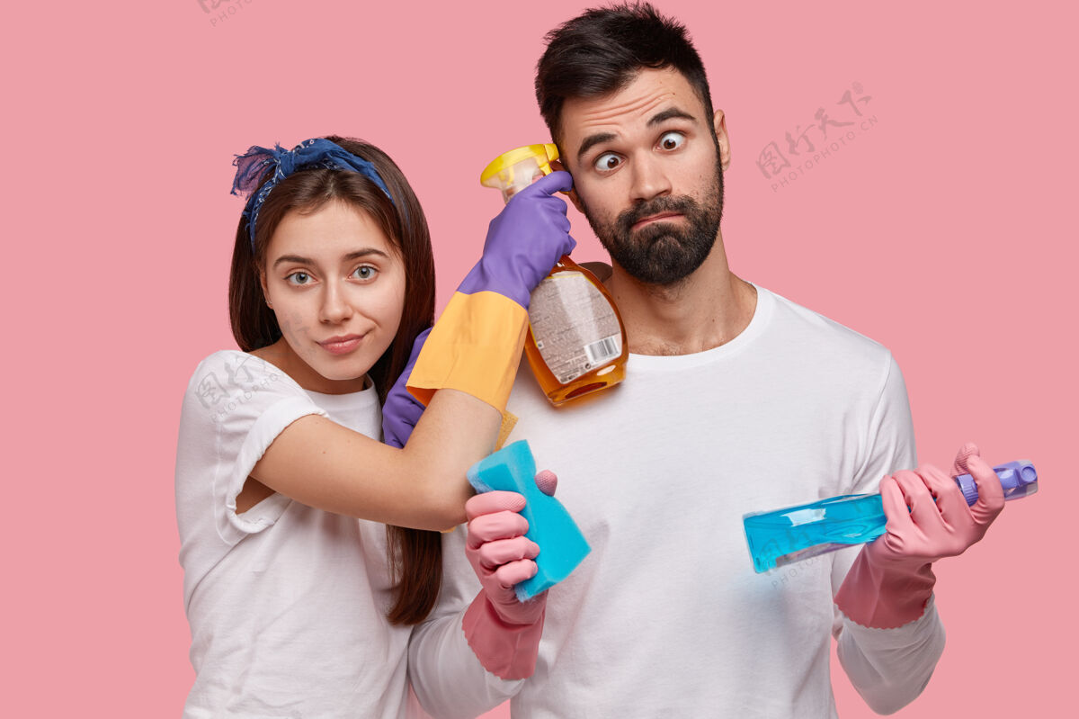 男朋友有趣的没刮胡子的男人交叉眼睛 帮妻子打扫房子 一起工作 做家务 用洗涤剂 海绵杂务搞笑墙