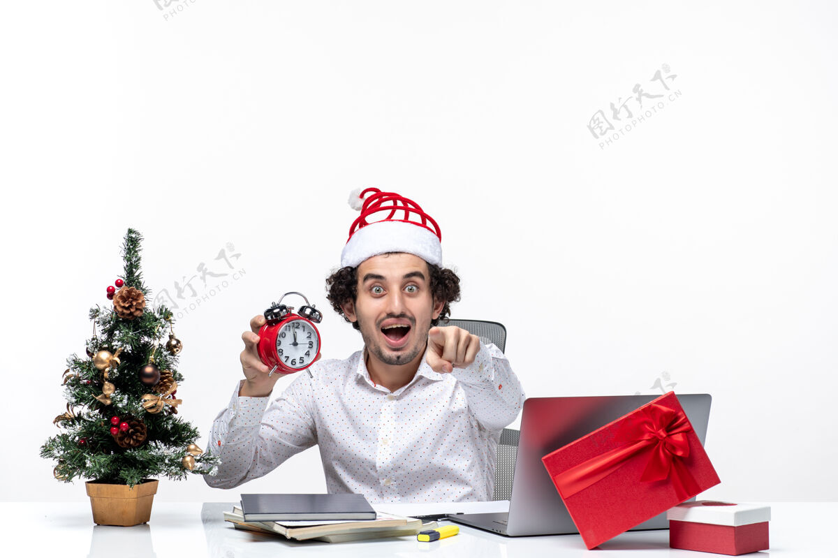 时钟年轻兴奋的商人戴着圣诞老人的帽子 在白色的背景下独自坐在办公室里看时钟圣诞老人帽子兴奋