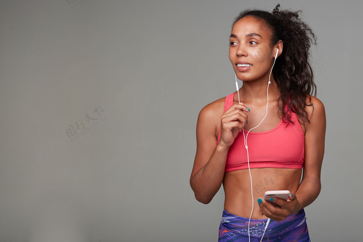 锻炼迷人的年轻棕色眼睛 皮肤黝黑 运动型的女士 早上锻炼前戴着耳机听音乐 站着耳机音乐粉色