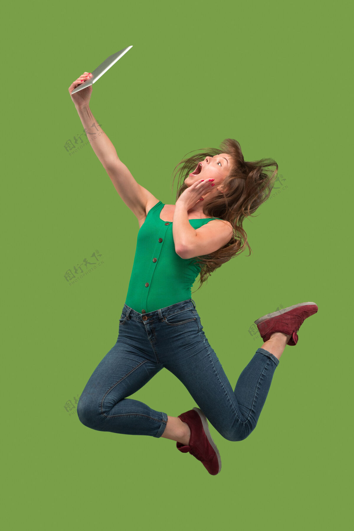 成功使用笔记本电脑或平板电脑小玩意在绿色工作室背景上跳跃的年轻女子在运动或运动中奔跑的女孩人类情感和面部表情概念现代生活中的小玩意手机女性服装