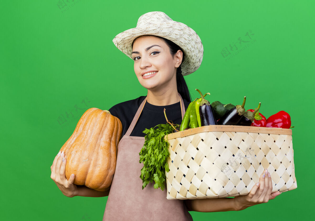 抱着年轻漂亮的女园丁 围着围裙 戴着帽子 手里拿着装满蔬菜和南瓜的箱子 满脸笑容绿色漂亮微笑