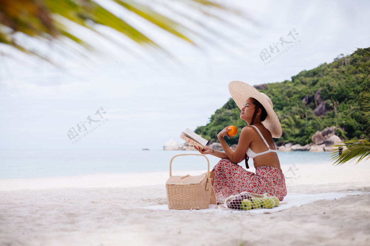迷人一个年轻女子戴着草帽 穿着白色针织衣服坐在沙滩地毯上女性美丽野餐