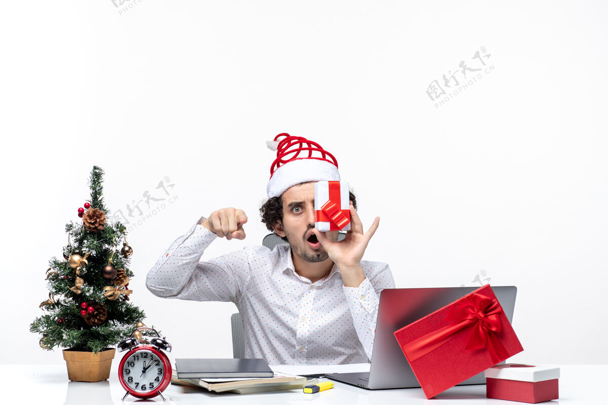 生意圣诞节心情震惊兴奋的商人与圣诞老人的帽子举起他的礼物 他的脸 并指出白色背景上的东西礼物人喜剧演员