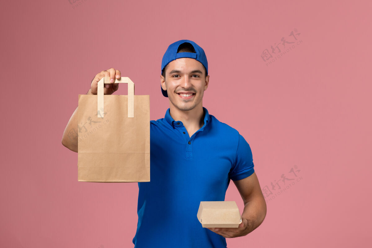 人正面图：身穿蓝色制服和披风的年轻男性快递员 手上拿着快递包裹 站在粉红色的墙上快乐送货视图